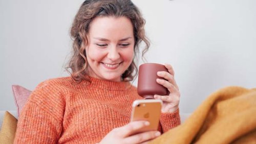 Smilende ung kvinde sidder i sofaen med kaffekop og telefon i hænderne.